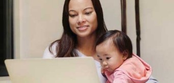 An individual on their laptop while holding a child.