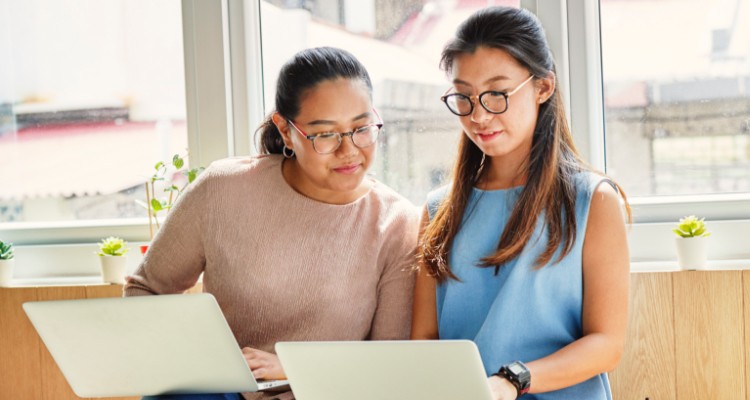 Two young women wearing glasses discuss business with their laptops open